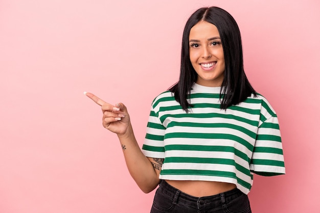 Jonge blanke vrouw met één arm geïsoleerd op roze achtergrond glimlachend en opzij wijzend, iets tonend op lege ruimte.