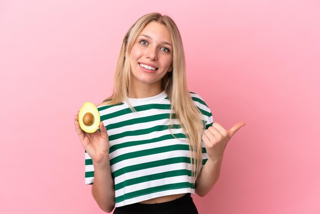 Jonge blanke vrouw met een avocado geïsoleerd op een roze achtergrond die naar de zijkant wijst om een product te presenteren