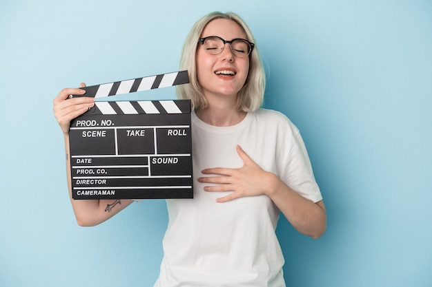 Jonge blanke vrouw met een Filmklapper geïsoleerd op blauwe achtergrond lacht hardop terwijl ze de hand op de borst houdt.