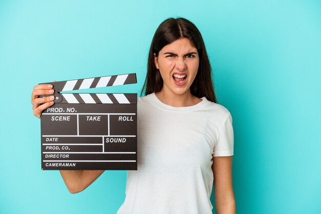 Jonge blanke vrouw met een Filmklapper geïsoleerd op een blauwe achtergrond schreeuwend erg boos en agressief.