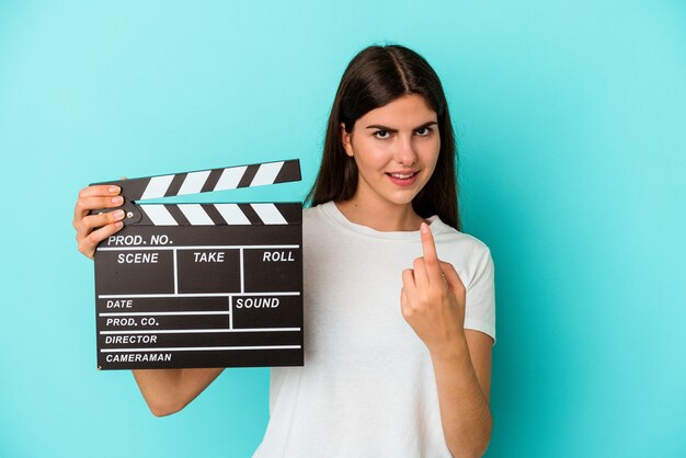 Jonge blanke vrouw met een Filmklapper geïsoleerd op een blauwe achtergrond, wijzend met de vinger naar je alsof uitnodigend dichterbij komt.