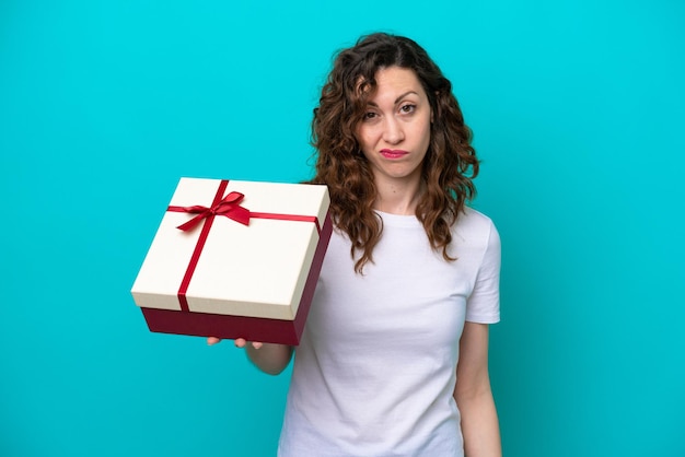 Jonge blanke vrouw met een geschenk geïsoleerd op een blauwe achtergrond met droevige uitdrukking