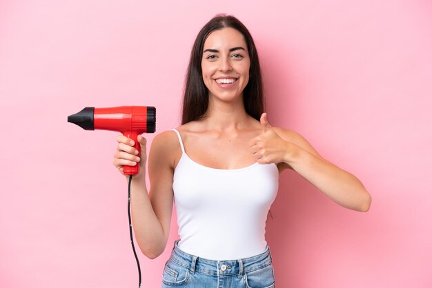 Jonge blanke vrouw met een haardroger geïsoleerd op roze achtergrond met duimen omhoog omdat er iets goeds is gebeurd