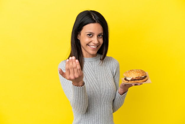 Jonge blanke vrouw met een hamburger geïsoleerd op een gele achtergrond die uitnodigt om met de hand te komen. Blij dat je gekomen bent