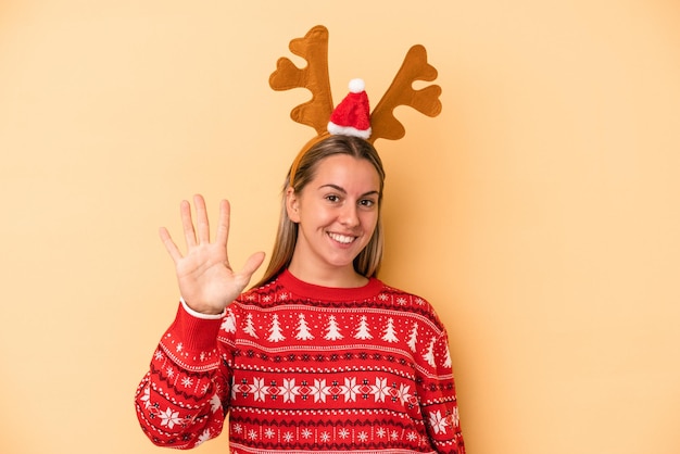 Jonge blanke vrouw met een kerst rendier hoed geïsoleerd op een gele achtergrond glimlachend vrolijk met nummer vijf met vingers.
