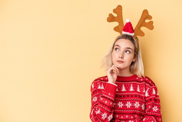 Foto jonge blanke vrouw met een kerst rendier hoed geïsoleerd op een gele achtergrond zijwaarts kijkend met twijfelachtige en sceptische uitdrukking.