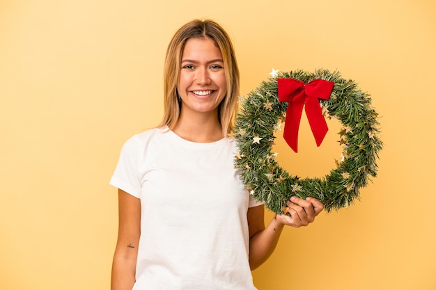 Jonge blanke vrouw met een kerstkrans geïsoleerd op gele achtergrond gelukkig, glimlachend en vrolijk.