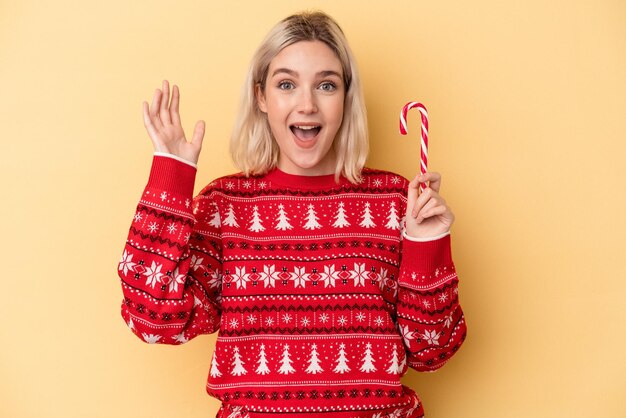 Jonge blanke vrouw met een kerststok geïsoleerd op gele achtergrond die een aangename verrassing ontvangt, opgewonden en handen opsteekt.