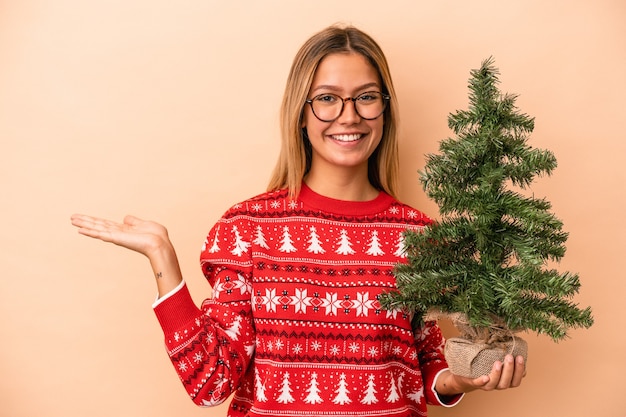 Jonge blanke vrouw met een kleine kerstboom geïsoleerd op een beige achtergrond met een kopie ruimte op een palm en met een andere hand op de taille.