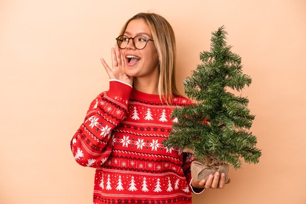 Jonge blanke vrouw met een kleine kerstboom geïsoleerd op een beige achtergrond, schreeuwend en met palm in de buurt van geopende mond.