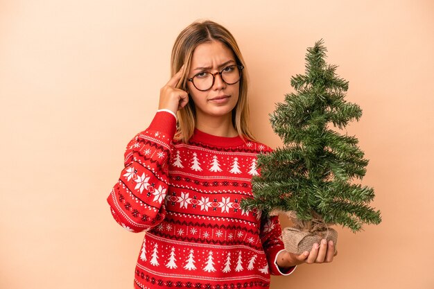 Jonge blanke vrouw met een kleine kerstboom geïsoleerd op een beige achtergrond wijzende tempel met vinger, denken, gericht op een taak.