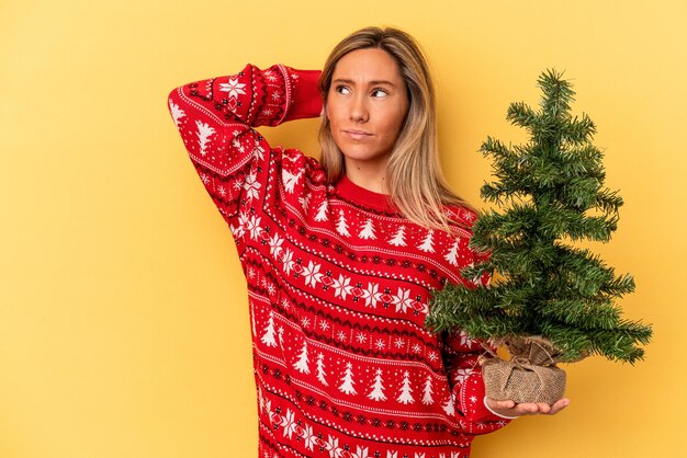 Jonge blanke vrouw met een kleine kerstboom geïsoleerd op een gele achtergrond die de achterkant van het hoofd aanraakt, denkt en een keuze maakt.