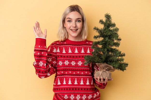 Jonge blanke vrouw met een kleine kerstboom geïsoleerd op een gele achtergrond die een aangename verrassing ontvangt, opgewonden en handen opsteekt.