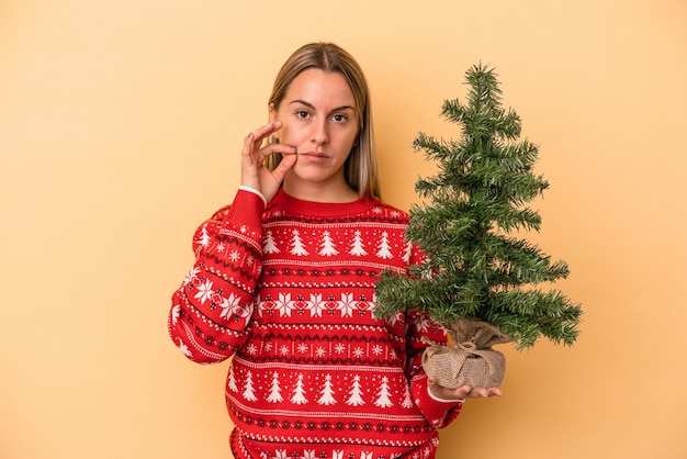 Jonge blanke vrouw met een kleine kerstboom geïsoleerd op een gele achtergrond met vingers op de lippen die een geheim bewaren.