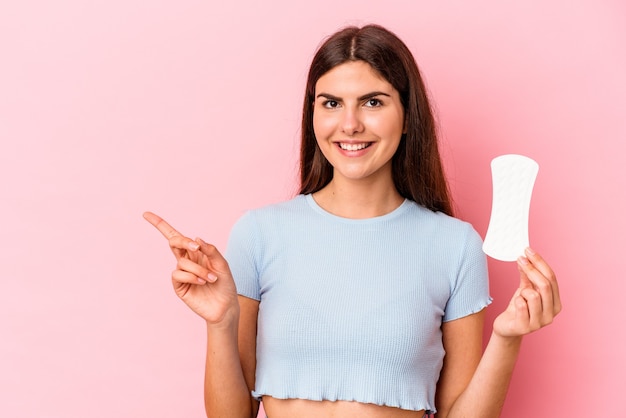 Jonge blanke vrouw met een kompres geïsoleerd op roze achtergrond glimlachend en opzij wijzend, iets tonend op lege ruimte.