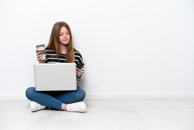 Jonge blanke vrouw met een laptop zittend op de vloer geïsoleerd op een witte achtergrond met koffie om mee te nemen en een mobiel