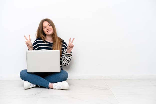 Jonge blanke vrouw met een laptop zittend op de vloer geïsoleerd op een witte achtergrond met overwinningsteken met beide handen