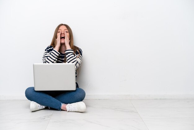 Jonge blanke vrouw met een laptop zittend op de vloer geïsoleerd op een witte achtergrond schreeuwen en iets aankondigen