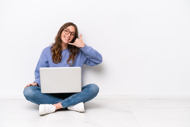 Jonge blanke vrouw met een laptop zittend op de vloer geïsoleerd op een witte achtergrond telefoon gebaar maken Bel me terug teken