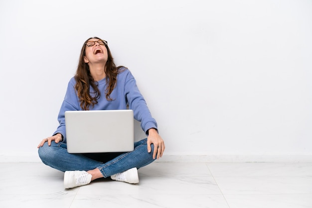 Jonge blanke vrouw met een laptop zittend op de vloer geïsoleerd op witte achtergrond lachen
