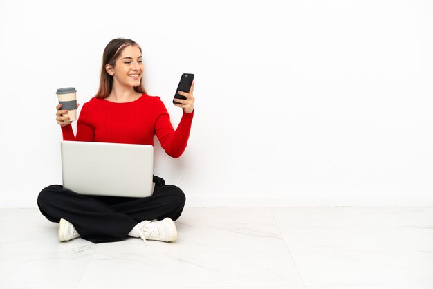 Jonge blanke vrouw met een laptop zittend op de vloer met koffie om mee te nemen en een mobiel