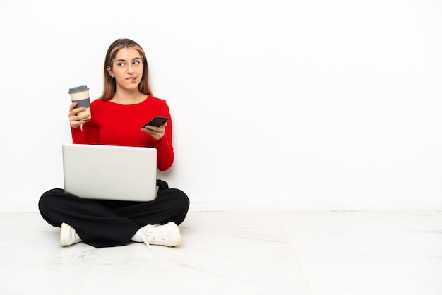 Jonge blanke vrouw met een laptop zittend op de vloer met koffie