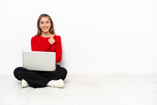 Jonge blanke vrouw met een laptop zittend op de vloer wijzend naar de zijkant om een product te presenteren