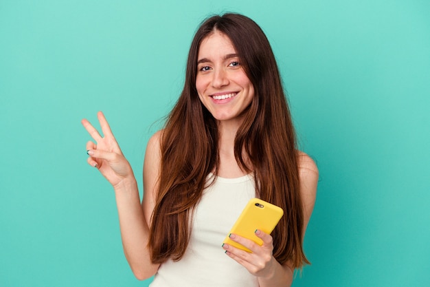 Jonge blanke vrouw met een mobiele telefoon geïsoleerd op blauwe achtergrond blij en zorgeloos met een vredessymbool met vingers.