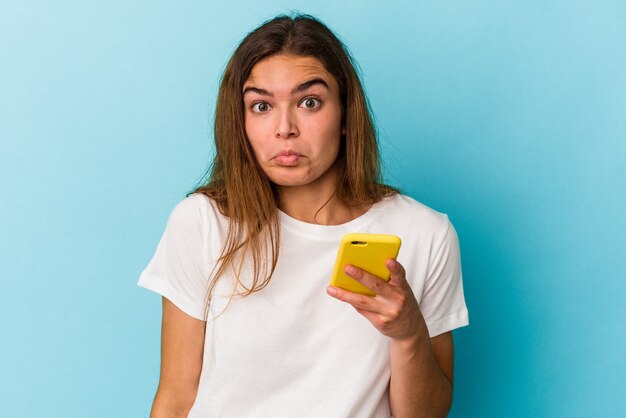 Jonge blanke vrouw met een mobiele telefoon geïsoleerd op blauwe achtergrond haalt schouders op en open ogen verward.