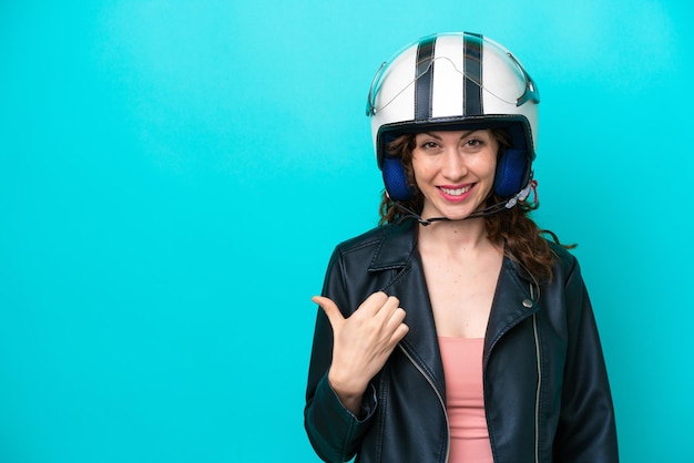 Jonge blanke vrouw met een motorhelm geïsoleerd op een blauwe achtergrond die naar de zijkant wijst om een product te presenteren