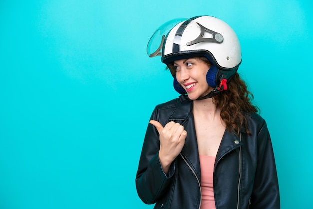 Jonge blanke vrouw met een motorhelm geïsoleerd op een blauwe achtergrond die naar de zijkant wijst om een product te presenteren