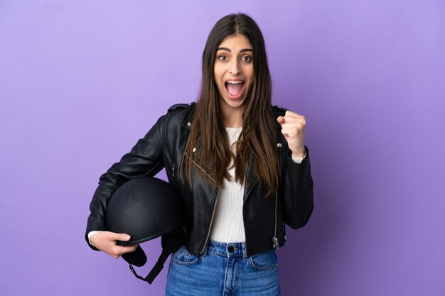 Jonge blanke vrouw met een motorhelm geïsoleerd op een paarse achtergrond die een overwinning viert in winnaarspositie