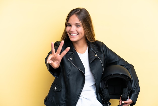 Jonge blanke vrouw met een motorhelm geïsoleerd op gele achtergrond gelukkig en drie tellen met vingers
