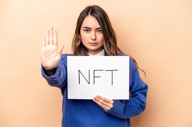 Jonge blanke vrouw met een NFT-plakkaat geïsoleerd op een beige achtergrond, staande met uitgestrekte hand met stopbord dat je verhindert