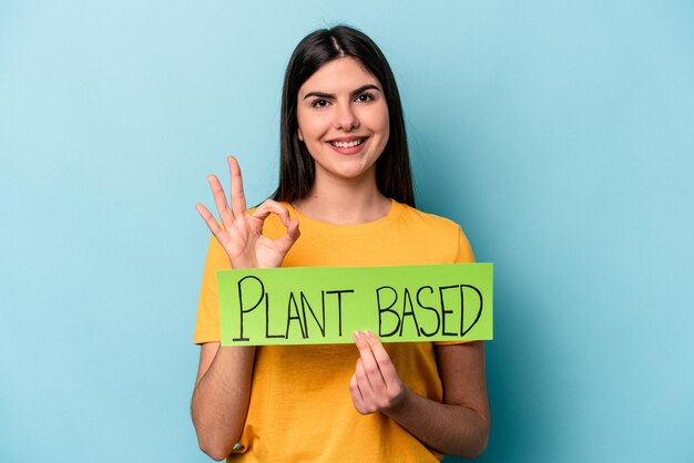 Jonge blanke vrouw met een op planten gebaseerd bordje geïsoleerd op een blauwe achtergrond, vrolijk en zelfverzekerd met een goed gebaar