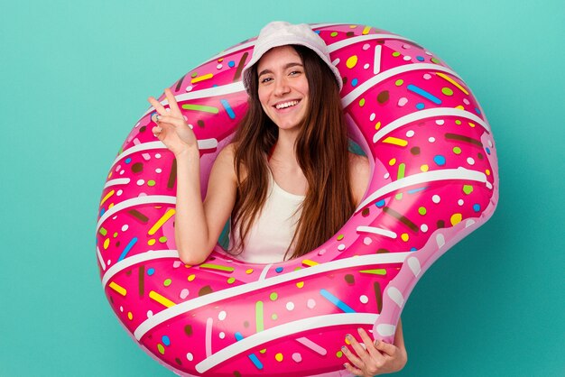 Jonge blanke vrouw met een opblaasbare donut geïsoleerd op blauwe achtergrond blij en zorgeloos met een vredessymbool met vingers.