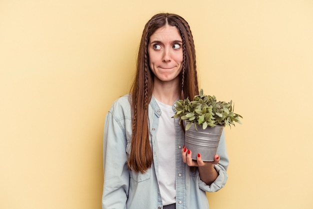 Jonge blanke vrouw met een plant geïsoleerd op een gele achtergrond verward, voelt zich twijfelachtig en onzeker.