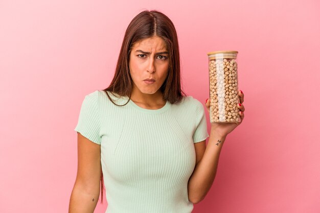 Jonge blanke vrouw met een pot kikkererwten geïsoleerd op roze achtergrond haalt schouders op en open ogen verward.