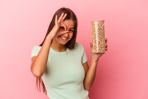 Jonge blanke vrouw met een pot kikkererwten geïsoleerd op roze achtergrond opgewonden houden ok gebaar in de gaten.