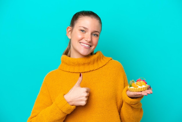 Jonge blanke vrouw met een taartje geïsoleerd op een blauwe achtergrond met duimen omhoog omdat er iets goeds is gebeurd