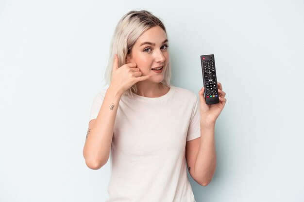 Jonge blanke vrouw met een tv-controller geïsoleerd op een blauwe achtergrond met een mobiel telefoongesprek gebaar met vingers.