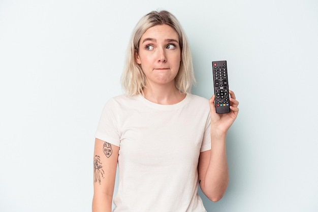 Jonge blanke vrouw met een tv-controller geïsoleerd op een blauwe achtergrond verward, voelt zich twijfelachtig en onzeker.