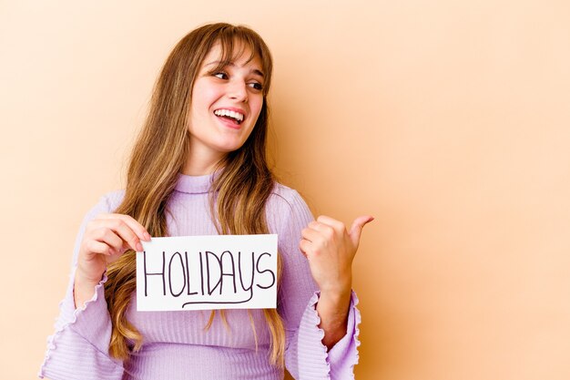 Jonge blanke vrouw met een vakantie-aanplakbiljet geïsoleerde punten met duimvinger weg, lachen en zorgeloos