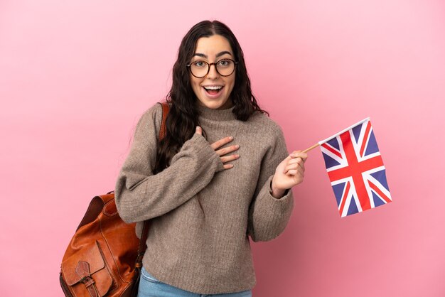 Jonge blanke vrouw met een vlag van het Verenigd Koninkrijk geïsoleerd op roze achtergrond verrast en geschokt terwijl ze naar rechts kijkt