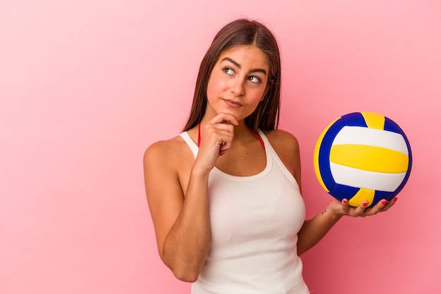 Jonge blanke vrouw met een volleybal bal geïsoleerd op roze achtergrond zijwaarts kijkend met twijfelachtige en sceptische uitdrukking.