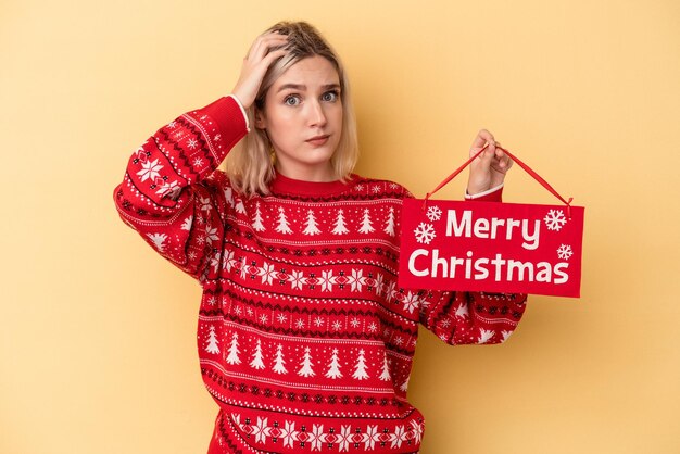 Jonge blanke vrouw met een vrolijk kerstbordje geïsoleerd op een gele achtergrond die geschokt is, herinnert ze zich een belangrijke vergadering.