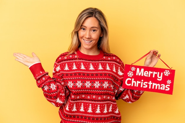 Jonge blanke vrouw met een vrolijk kerstfeestje geïsoleerd op een gele achtergrond met een kopie ruimte op een palm en met een andere hand op de taille.