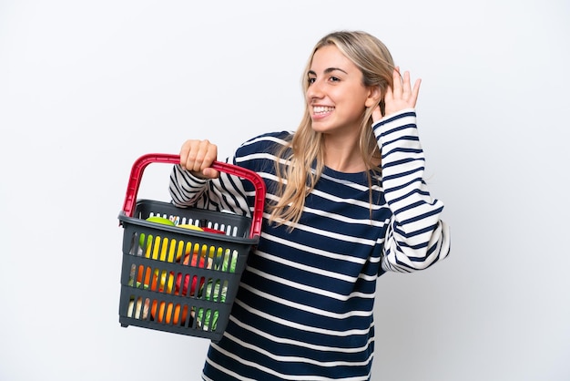 Jonge blanke vrouw met een winkelmandje vol eten geïsoleerd op een witte achtergrond luisterend naar iets door de hand op het oor te leggen