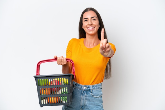 Jonge blanke vrouw met een winkelmandje vol voedsel geïsoleerd op een witte achtergrond die een vinger toont en opheft