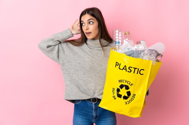 Jonge blanke vrouw met een zak vol met plastic flessen om te recyclen geïsoleerd op roze achtergrond luisteren naar iets door hand op het oor te leggen